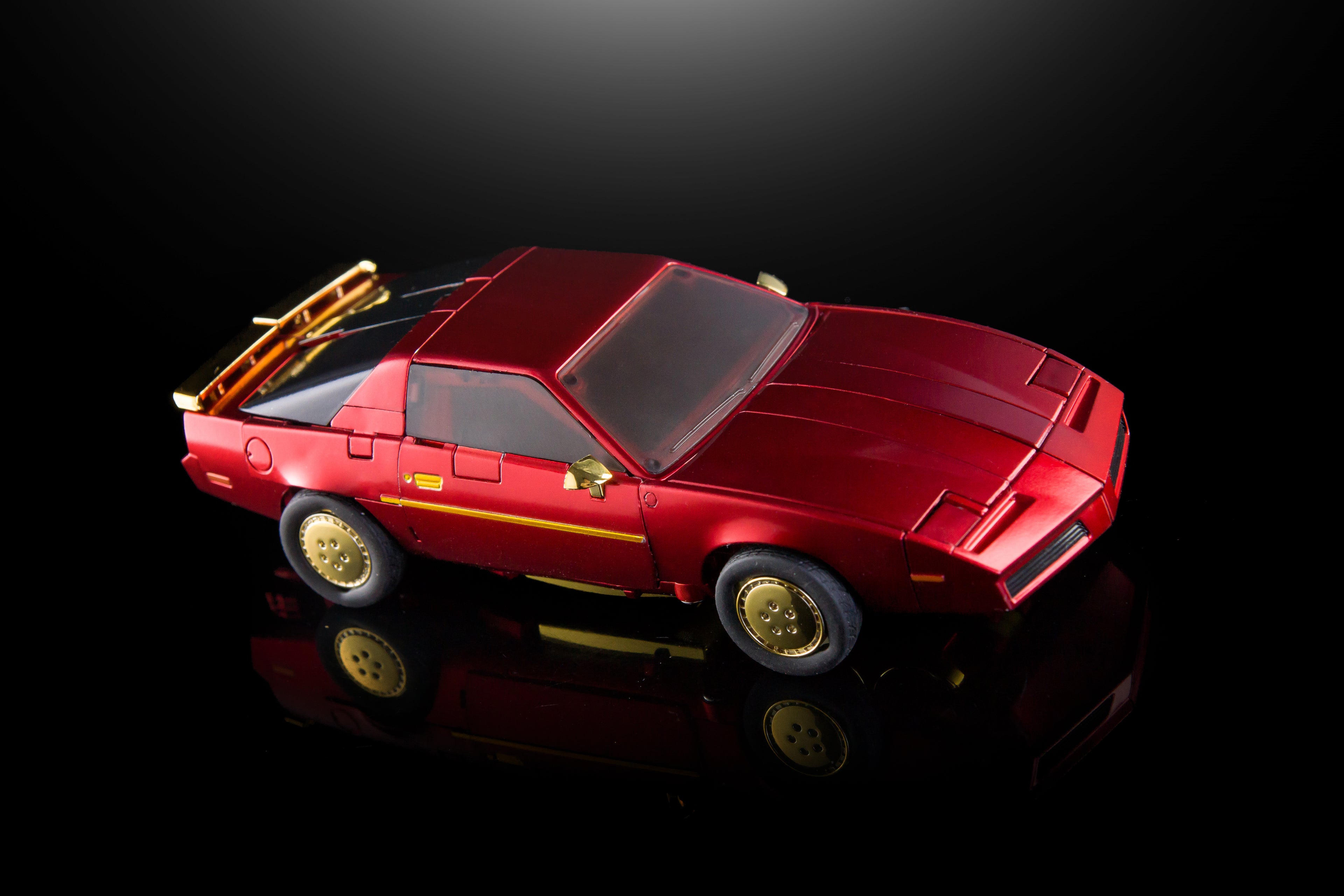 A red model car with gold wheels and trim, displayed on a reflective black surface against a dark background.
