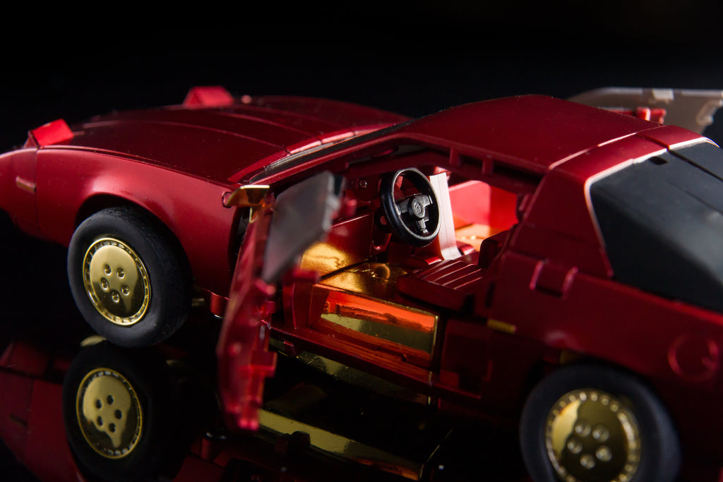 A red toy sports car with gold wheels and an open door revealing a detailed gold interior, photographed on a dark reflective surface against a black background.