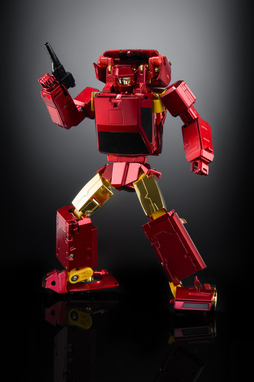 A red and gold metal action figure or robot toy posed in a dynamic stance on a dark reflective studio background, holding a black object in one raised hand.